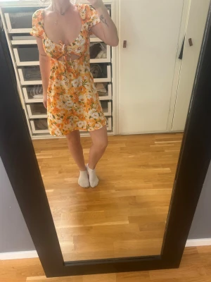 Blommig kortklänning med knyt - Söt kortklänning med blommigt mönster i orange, vit och gul. Klänningen har korta puffärmar, v-ringning och knyt framtill. Tillverkad i ett lätt och luftigt material, perfekt för varma dagar. Den passar även Small!