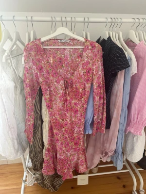 Blommig rosa klänning från Zara - Söt kort klänning från Zara med blommigt mönster i rosa och vita nyanser. Klänningen har v-ringning med snörning framtill, volang nertill och långa ärmar. Perfekt för sommarens alla tillfällen.