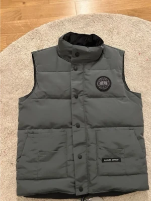Grå dunväst från Canada Goose, möts bara upp i Stockholm  - Säljer en grå dunväst från Canada Goose med hög krage och två stora fickor framtill. Västen har ett svart Canada Goose-märke på bröstet och är tillverkad i slitstarkt material med dunfyllning. Perfekt för kalla dagar och snygg att ha över en hoodie.