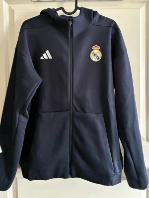 Mörkblå Real Madrid hoodie Adidas - Snygg mörkblå hoodie från Adidas med Real Madrid-logga på bröstet och Adidas-logga i vitt. Dragkedja framtill, två fickor och huva. Perfekt för dig som älskar fotboll och vill ha en sportig look. Tillverkad i mjukt material för extra komfort.