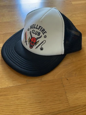 Hellfire Club keps svart/vit - Cool keps med Hellfire Club-motiv från Stranger Things. Framsidan är vit med tryck av demonhuvud, eld och vapen, medan baksidan är svart mesh för extra ventilation. Perfekt för dig som gillar retrostil och popkultur.