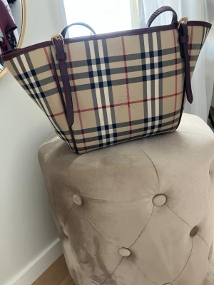 Burberry rutig handväska i canvas - Säljer min fina och klassiska handväska från Burberry i beige canvas med ikoniskt rutmönster. Insidan är mörkt röd/brun. Mycket väl omhändertagen och utan några egentliga tecken på användning. Perfekt väska för sommaren för att enkelt bära med sig det viktigaste. Längden på handtagen kan justeras. Köpt direkt från butiken. Kvitto och mer information kan ges. 
