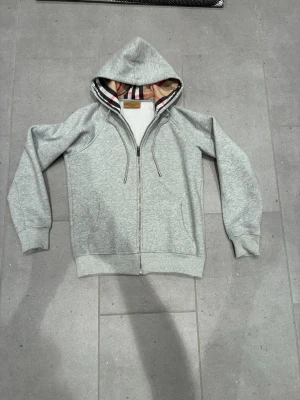 Grå burberry hoodie  - Säljer en grå hoodie från Burberry med dragkedja och klassiskt rutigt foder i huvan. Hoodien har broderad logga på bröstet, ribbade muddar och snören i huvan. Perfekt för dig som vill ha en stilren och exklusiv look.