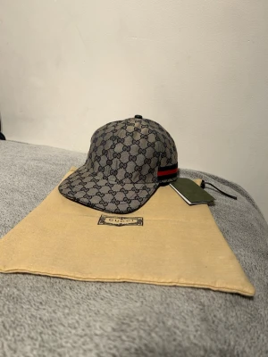 Gucci keps med GG-mönster - Snygg keps från Gucci i beige och mörkblått med klassiskt GG-monogram över hela kepsen. På sidan finns en röd och marinblå rand. Justerbar rem baktill och böjd skärm. Tillverkad i canvasmaterial. Perfekt för dig som vill ha en exklusiv och trendig look.