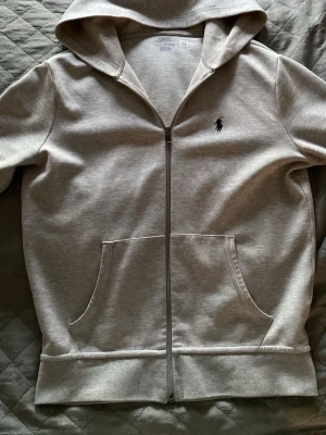 Grå zip hoodie från Polo Ralph Lauren - Snygg grå hoodie med dragkedja från Polo Ralph Lauren. Klassisk design med huva, två stora fickor framtill och den ikoniska mörkblå polospelaren broderad på bröstet. Tillverkad i mjukt bomullsmaterial som känns skönt mot huden.