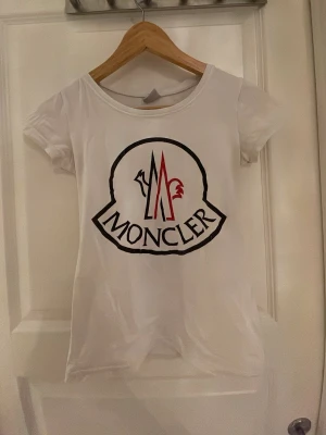 Vit Moncler t-shirt - Säljer en vit t-shirt från Moncler med stort logotryck i svart och rött på framsidan. Den är i stl s men passar xs okså den är tajt så den ger fin figur och är snygg till jeans och den är i 100% bomull 