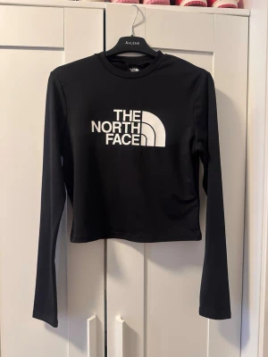 Svart långärmad crop top The North Face - Snygg svart långärmad crop top från The North Face med stor vit logga framtill. Tröjan har rund halsringning och är gjord i ett stretchigt material som sitter tajt. Perfekt för en sportig och trendig look.