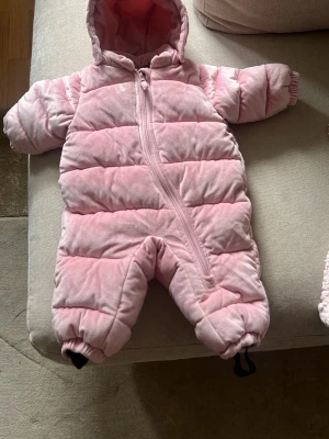 Rosa vadderad overall med huva - Mysig rosa overall med huva och dragkedja framtill. Overallen är vadderad och har resår vid ärm- och benslut samt praktiska fothällor. Perfekt för kalla dagar och enkel att ta på tack vare den långa dragkedjan.jag kan gå ner i pris