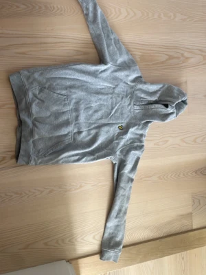Grå hoodie från Lyle & Scott - Säljer en grå hoodie från Lyle & Scott med klassisk gul logga på bröstet. Hoodien har huva med dragsko och en stor magficka framtill. Tillverkad i mjukt sweatshirtmaterial och har ribbade muddar vid ärmar och nederkant.