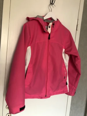 Rosa vindjacka med huva från Everest - Snygg rosa vindjacka/seglarjacka från Everest med vita detaljer på sidorna och justerbar huva. Jackan har dragkedja framtill, två fickor och kardborre vid ärmsluten. Perfekt för blåsiga dagar och enkel att matcha med olika outfits. Vind- och vattenresistent, fantastisk kvalitet av det tjockare slaget, andningsbarinsida, se bilder.
