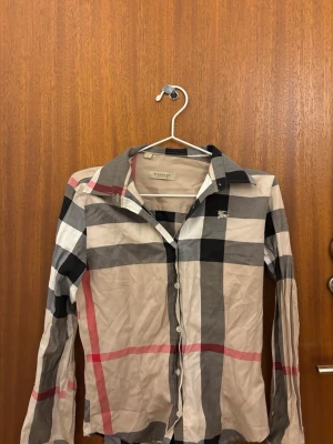 Rutig skjorta från Burberry London(vet inte om de är äkta) - Klassisk rutig skjorta från Burberry London i beige, svart, vit och röd. Skjortan har långa ärmar, knappar framtill och den ikoniska Burberry-loggan broderad på bröstet. Perfekt för dig som gillar tidlös och stilren design.