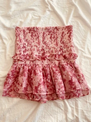 Rosa volangkjol med smock - Supersöt kort kjol i rosa och vitt med smock upptill och volangdetaljer. Kjolen har ett blommigt mönster och är gjord i ett lätt, luftigt material som passar perfekt till varmare dagar. Elastisk midja för skön passform och extra volym i kjolen. Skriv för fler bilder🥰