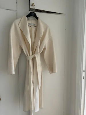Krämvit kappa från Zara med skärp - Stilren krämvit kappa från Zara med breda slag och knytskärp i midjan. Kappan har en rak passform och är tillverkad i ett mjukt tyg som ger en lyxig känsla. Perfekt för att lyfta vilken outfit som helst.