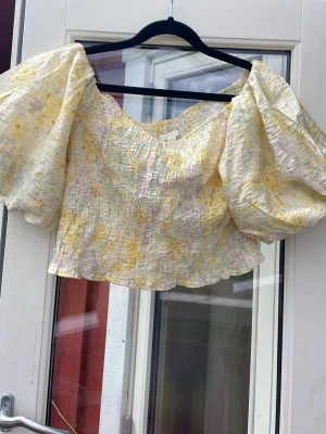 Gul blommig topp med puffärmar - Säljer en söt croppad topp med stora puffärmar och smockad struktur. Toppen har ett blommigt mönster i gult, vitt och ljuslila. Perfekt för dig som gillar romantisk och trendig stil. Materialet känns lätt och luftigt.