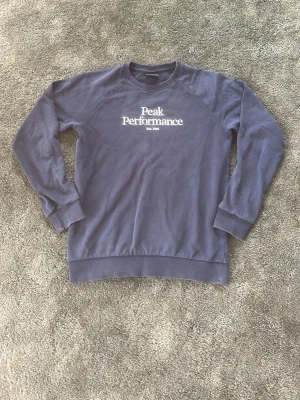 Blå sweatshirt från Peak Performance - Mörkblå sweatshirt från Peak Performance med vit broderad logga på bröstet. Klassisk rund halsringning, ribbade muddar vid ärmslut och nederkant. Perfekt för en avslappnad och sportig stil.