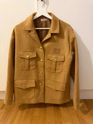  jacka  från Miu Miu - Stilren jacka från Miu Miu med fyra stora fickor framtill och guldiga knappar. Overshirten har klassisk krage och broderad logga på bröstet. Tillverkad i ett mjukt material som ger en lyxig känsla och passar perfekt till lager-på-lager. Storleken är S men passar S-M eftersom ganska oversize