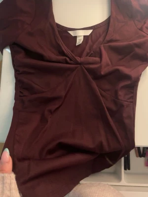 Vinröd v-ringad topp H&M - Snygg vinröd topp från H&M med v-ringning och lätt rynkdetalj framtill. Modellen har korta ärmar och är gjord i ett mjukt, stretchigt och glittrigt material som sitter bekvämt på kroppen. Perfekt att styla med jeans eller kjol. 