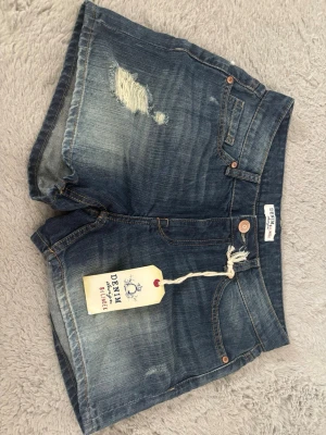 Blå denimshorts från Denim & Supply - Snygga blå jeansshorts från Denim & Supply med slitna detaljer och klassisk femficksdesign. Shortsen har normal passform, knappgylf och råa kanter för en avslappnad look. Perfekta till sommaren och lätta att matcha med olika stilar. Lindex betyg