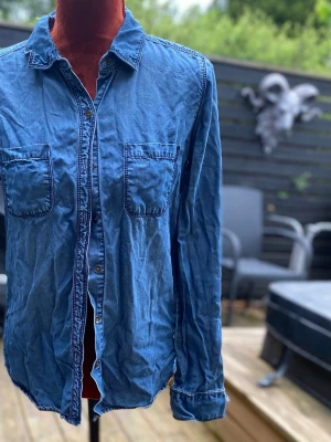 Blå jeansskjorta med fickor S - Skjorta i 100% Lycocell med långa ärmar och krage. Skjortan har två bröstfickor med lock och knäppning framtill. Ärmsluten har knappar för justering. En avslappnad passform som passar bra för vardagsbruk. Längd: 67 cm Bystvidd: 43 cm  Inga bilder på mig! Katter finns i hemmet!