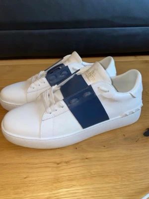 Valentino vita sneakers med blå detalj bytes mot andra skor - Snygga vita sneakers från Valentino med en bred blå remsa över snörningen och små nitar på hälen. Tillverkade i slätt läder med klassisk låg siluett och vita skosnören. Perfekta för dig som gillar stilrena men ändå unika skor. Gammal bild använda 3 gånger ser helt nya ut kan skicka fler bilder i privat om man är intresserad 