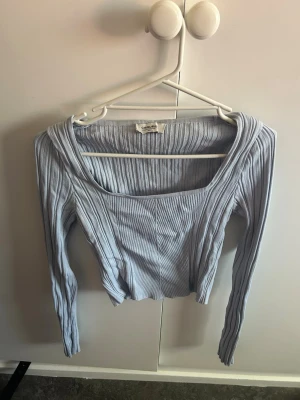 Ljusblå ribbad topp från CHQUELLE - Trendig ljusblå ribbad topp från Gina Tricot med fyrkantig ringning och långa ärmar. Toppen är croppad och har en slim passform som framhäver figuren. Perfekt att styla med höga jeans eller kjol för en cool look.