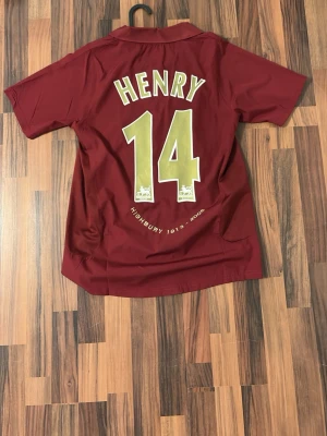 Arsenal 2005-6 retro tröja ( Henry ) - Säljer en ikonisk vinröd Arsenal matchtröja från Nike med Henry 14 på ryggen och klubbmärke samt O2-logga i guld på bröstet. Tröjan har korta ärmar, klassisk krage och är tillverkad i lätt, ventilerande material. Perfekt för fotbollsfans och samlare.