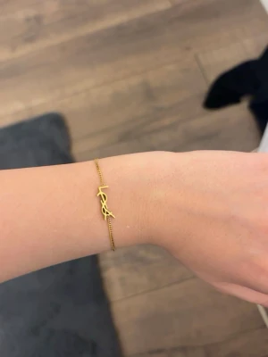 Guldigt armband från Saint Laurent - Snyggt och stilrent armband i guldton från Saint Laurent med den ikoniska YSL-loggan framtill. Tunt kedjearmband med justerbar längd och liten bricka med Saint Laurent Paris ingraverat vid låset. Perfekt accessoar för dig som gillar lyxiga detaljer.