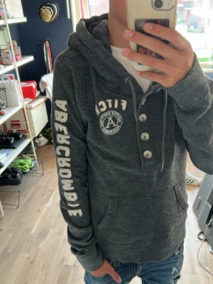 Mörkgrå hoodie från Abercrombie - Mörkgrå hoodie från Abercrombie & Fitch med stor logga på ärmen och tryck på bröstet. Hoodien har knappar framtill, snörning i huvan och en klassisk känguruficka. Perfekt för en chill och avslappnad stil.