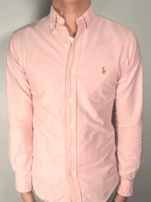 Ljusrosa skjorta från Ralph Lauren - Säljer en ljusrosa skjorta från Ralph Lauren i slim fit-modell. Skjortan är i bomull, har långa ärmar, klassisk krage med knappar och den ikoniska broderade loggan på bröstet. Perfekt för dig som gillar stilrena och snygga plagg.