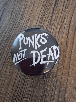 Punks Not Dead pinsknapp - Cool knapp med texten 'Punks Not Dead' i svart graffiti-stil. 4,4 cm. Perfekt accessoar för dig som gillar punk och vill sticka ut. Rund form och klassisk punkkänsla.