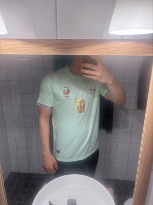 Frankrike Bortaställ VM 2026 Mbappé matchtröja - Ljusblå fotbollströja från Nike med Frankrikes landslagsemblem och nummer 10 på bröstet. På ryggen står det Mbappé och siffran 10. Tröjan har korta ärmar, rund hals och detaljer i mörkblått och rött.