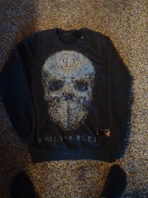 Svart tröja med dödskalle Philipp Plein - Cool svart tröja från Philipp Plein med stort grafiskt dödskalletryck på framsidan och märkets logga under. Tröjan har rund halsringning och ribbade muddar. Perfekt för dig som gillar streetwear och edgy stil.