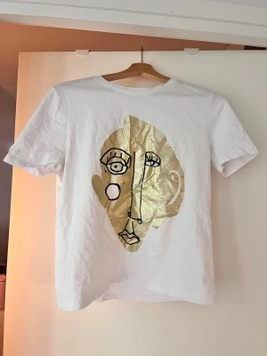 Vit t-shirt med guldtryck från Zara - Säljer en vit t-shirt från Zara med ett stort abstrakt ansikte i guld och svart tryck på framsidan. T-shirten har klassisk passform och korta ärmar. Perfekt för dig som gillar unika prints och vill sticka ut lite extra.
