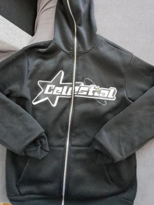 Svart hoodie Celestial Manfinity - Svart hoodie med dragkedja från Manfinity i storlek XS. På bröstet finns ett stort tryck med texten 'Celestial' och en stjärna. Hoodien har huva och två fickor framtill. Materialet är mjukt och känns fleece-liknande på insidan. Står XS men passar även S