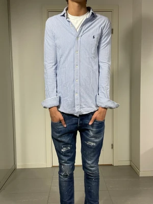 Blårandig skjorta från Ralph Lauren - Säljer en klassisk blårandig skjorta från Ralph Lauren med vit knappstängning och broderad logga på bröstet. Skjortan har lång ärm och normal passform, perfekt till jeans eller chinos. Tillverkad i mjuk bomull för en skön känsla.