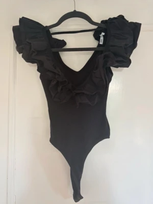 Svart body med volangärmar från Zara - Trendig svart body från zara med djup V-ringning och stora volangärmar som ger en statement-look. Tillverkad i stretchigt material med detaljer i chiffongliknande tyg på axlar och urringning. Perfekt för dig som vill sticka ut med din outfit.
