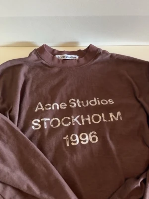 Acne Studios longsleeve - Cool brun sweatshirt från Acne Studios med tryck 'STOCKHOLM 1996' på bröstet. Tillverkad i 100% bomull och har en avslappnad passform med rund hals och långa ärmar. Perfekt för en trendig och avslappnad stil.