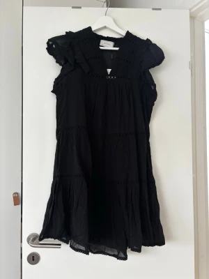 Svart volangklänning från Neo Noir - Supersöt svart klänning från Neo Noir med volangdetaljer och kort ärm. Klänningen har en luftig passform och fina broderade detaljer vid halsringningen. Perfekt för dig som gillar en romantisk och trendig stil. Nypris 899kr aldrig använd. 