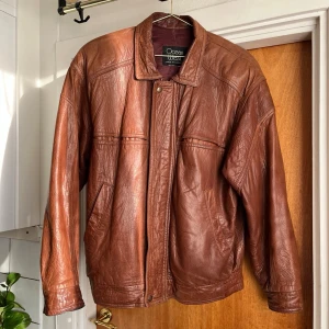 Brun skinnjacka från Ocean West - Snygg brun vintage skinnjacka från Ocean West med klassisk krage. Tidlös modell med avslappnad passform och diskreta sömdetaljer över bröstet. Perfekt för dig som gillar retrostil. Dragkjedan har tappat en del, men det har jag löst vid att festa en liten ring, så den fungerar som den skal