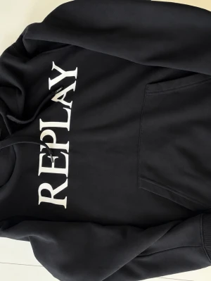 Svart hoodie från Replay - Snygg svart hoodie från Replay med stor vit logga över bröstet. Klassisk ficka framtill och justerbar huva med snören. Mjuk insida och ribbade muddar. Perfekt för dig som vill ha en stilren look. Pris 699