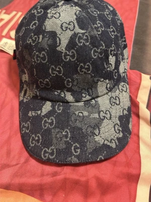 Gucci denimkeps med GG-mönster - Snygg keps från Gucci i blått denimtyg med det klassiska GG-monogrammet över hela kepsen. Kepsen har böjd skärm och knapp på toppen samt en liten grön-röd detalj baktill. Tillverkad i Italien och har en cool streetwear-vibe.
