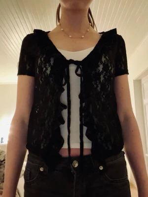 Svart spetsblus i kort modell - Svart topp i spets med kort ärm, volangdetaljer och knytning framtill. Lätt spetsmaterial som passar bra att styla med ett linne under. I fint skick 