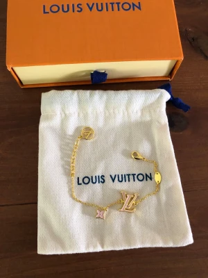 Louis Vuitton guldarmband med berlocker - Snyggt armband från Louis Vuitton i guldfärgad metall med berlocker i form av LV-logga, en rosa blomma och en rund detalj. Kedjan är tunn och elegant med karbinlås. Perfekt accessoar för dig som gillar exklusiv och trendig stil.