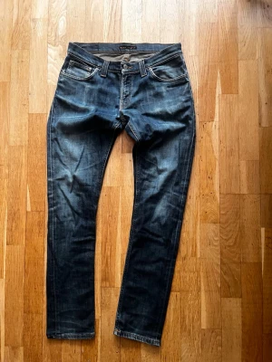 Mörkblå jeans från Nudie Jeans - Säljer ett par mörkblå jeans från Nudie Jeans med klassisk femficksmodell och raka ben. Jeansen har snygga slitningar och kontrastsömmar samt Nudie-logga på bakfickan. Tillverkade i denim med en skön passform.