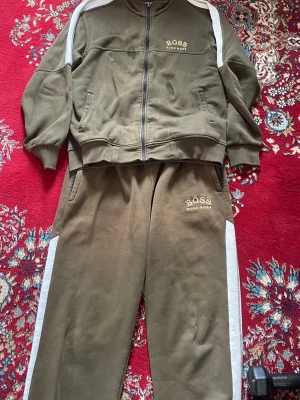 Olivgrönt loungewear-set från BOSS - Snyggt olivgrönt loungewear-set från BOSS Hugo Boss med vita sidopaneler på både byxor och tröja. Setet består av en ziptröja med fickor och matchande byxor med resår i midjan. Perfekt för chill dagar hemma eller när du vill vara bekväm men ändå se stilren ut.