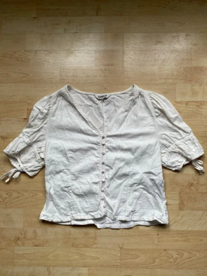 Vit blus med knappar från ONLY - Söt vit blus från ONLY med korta puffärmar och knytband vid ärmsluten. Blusen har v-ringning och dekorativa knappar framtill. Tillverkad i ett lätt och luftigt tyg, perfekt för en avslappnad och trendig look.
