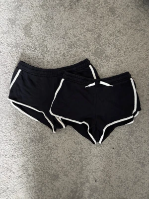 2 pack Svarta sportiga shorts med vit kant - Två par svarta shorts med vit kant och snörning i midjan. Klassisk sportig design med elastisk midja och kontrasterande detaljer längs sidorna och bensluten. Perfekta för en avslappnad och aktiv stil.