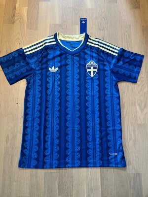 Blå Sverige fotbollströja Adidas - Säljer en blå fotbollströja från Adidas med Sveriges landslagsemblem på bröstet och gula detaljer på axlarna. Tröjan har ett unikt mönster i olika blå nyanser, korta ärmar och V-ringning med gul insida. Perfekt för match eller träning!
