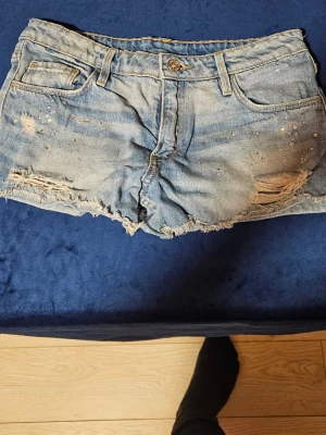 Ljusa jeansshorts med slitningar - Säljer ett par ljusblå jeansshorts med slitna detaljer, råa kanter och små nitar framtill. Klassisk femficksmodell med knapp och dragkedja. Perfekta för sommaren och ger en avslappnad vibe.