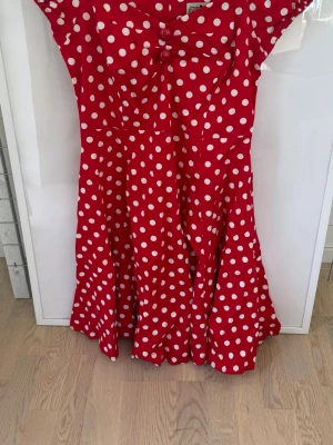 Röd prickig klänning med rosett - Säljer en röd klänning med vita prickar och en söt rosett framtill. Klänningen har kort ärm, markerad midja och utställd kjol som ger en klassisk retrovibe. Perfekt för dig som gillar färg och mönster! Endast använd en gång 
